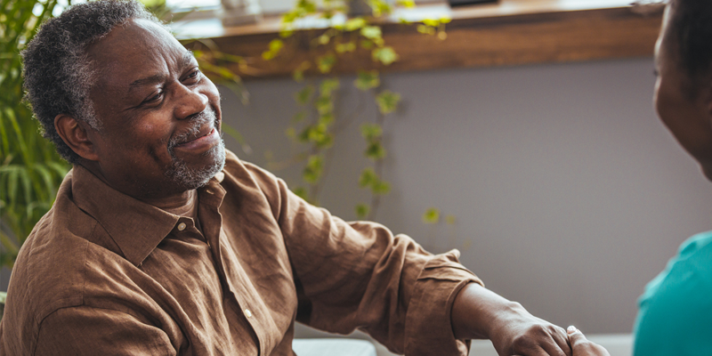 Elderly man sits on a couch holding hands with someone