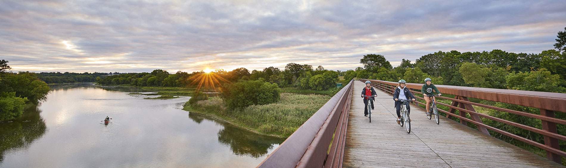 Three bikers ride their bikes on a trail along the Grand River during sunset