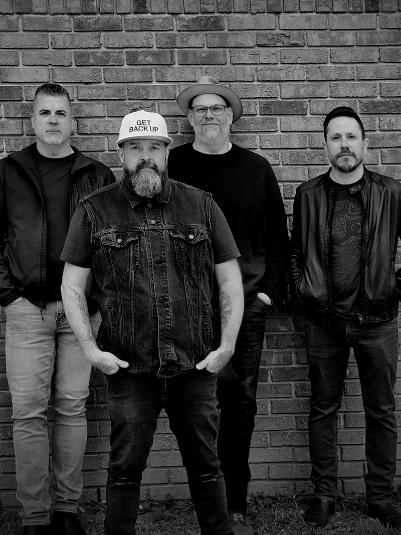A black and white photo of four men standing in front of a brick wall