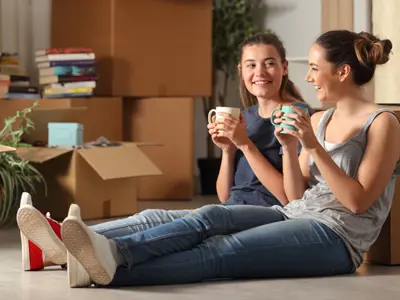 Two young girls sit on the floor of a living room surrounded by boxes